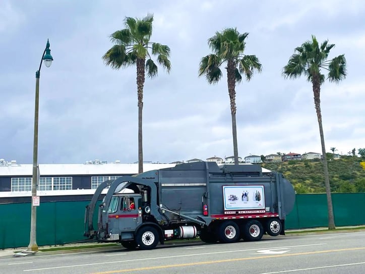 A blue garbage truck in the morning against a fence with palm trees in the back.