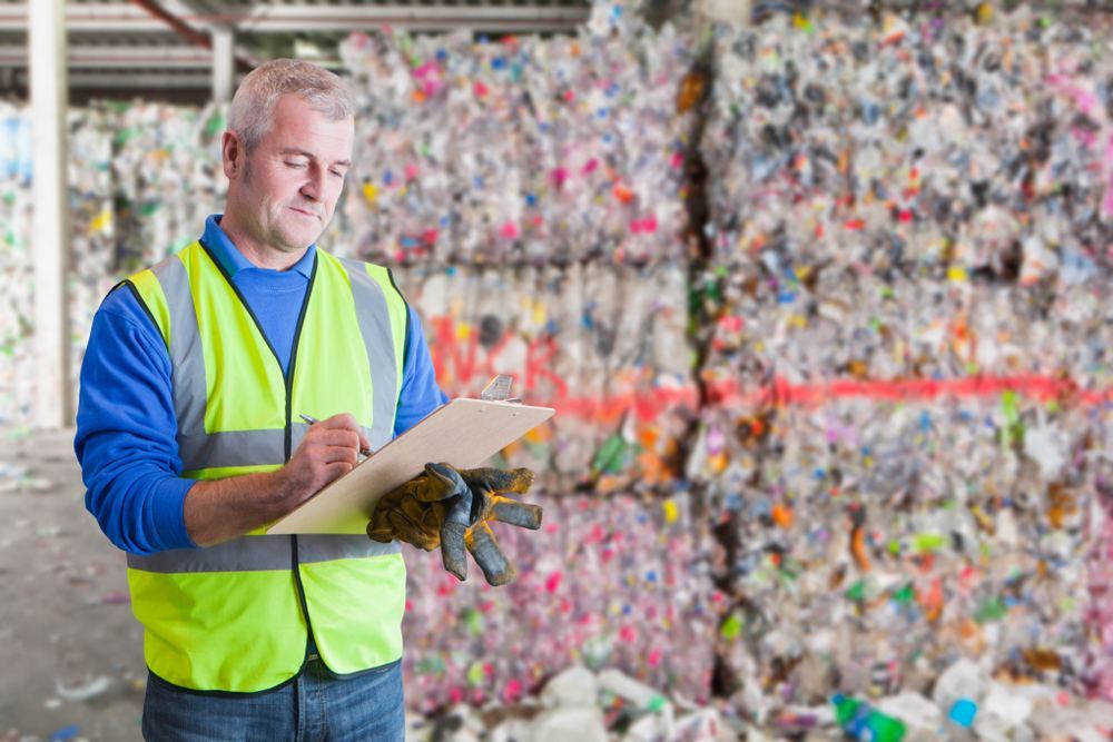 Facility worker uses a clipboard while inspecting stacked recycling bales at an industrial site.